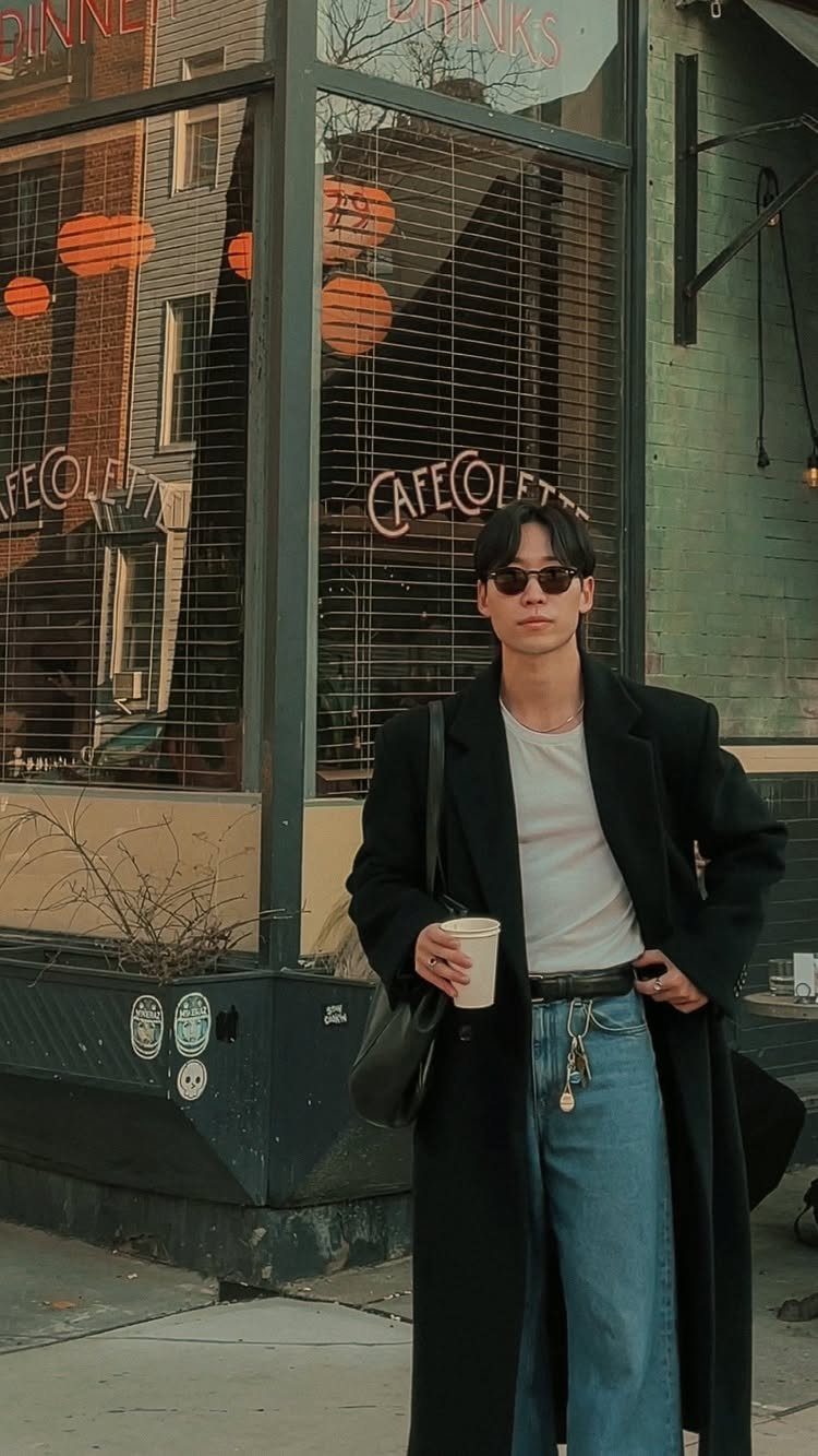 A man in a long black coat, white t-shirt, and blue jeans standing outdoors.
