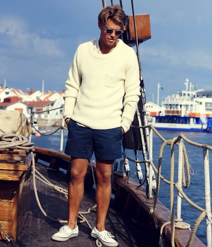 A young man in a white sweater and navy shorts posing on a wooden boat deck near a harbor.