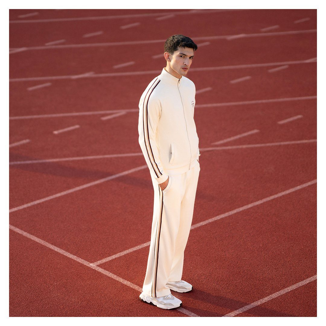Man in a cream track suit and white sneakers standing on a red running track.