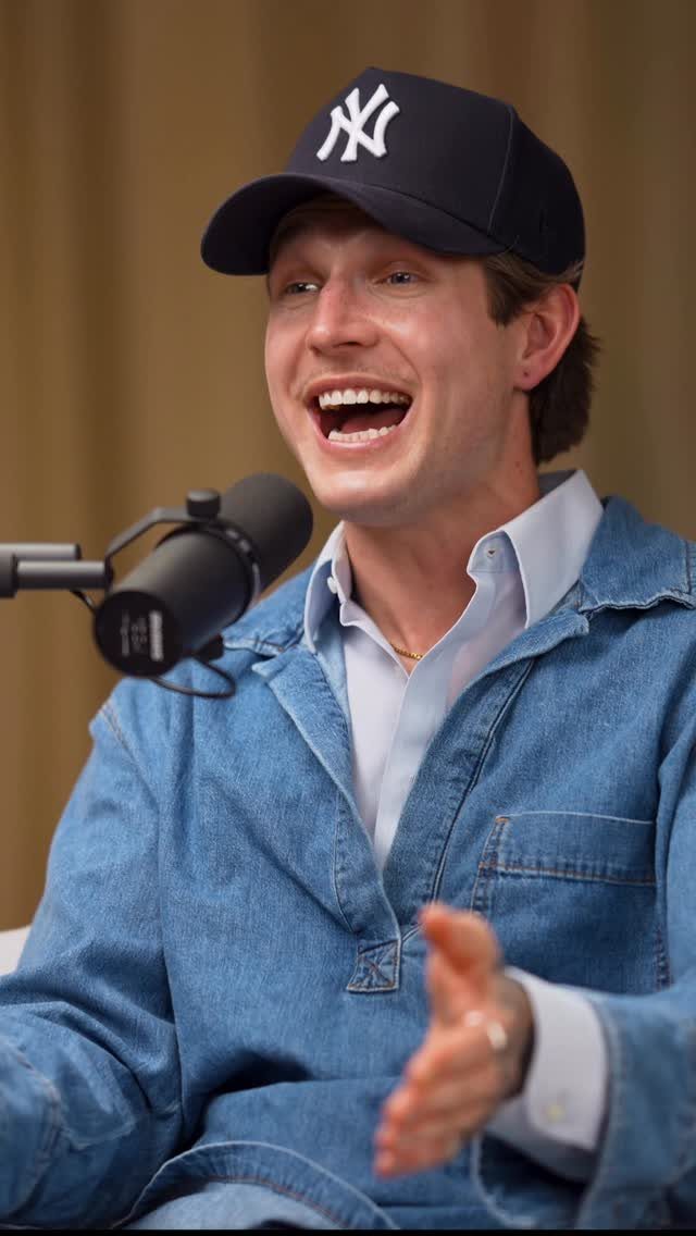 A man in a denim shirt and Yankees cap speaking into a microphone while laughing.