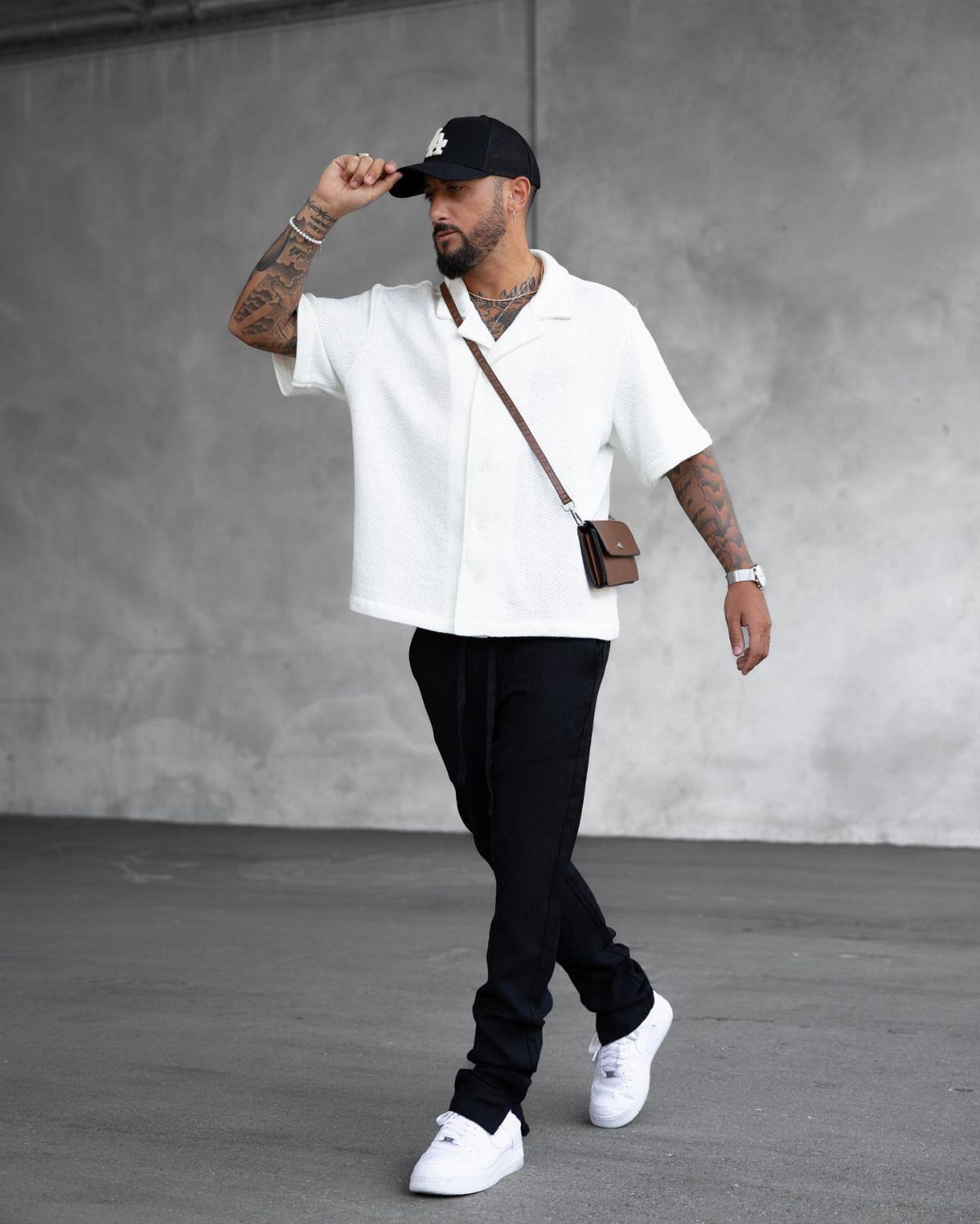 A man in a white shirt and black pants walking against a grey concrete wall.