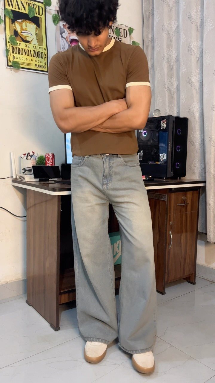 A young man in a brown t-shirt and wide jeans posing with crossed arms.