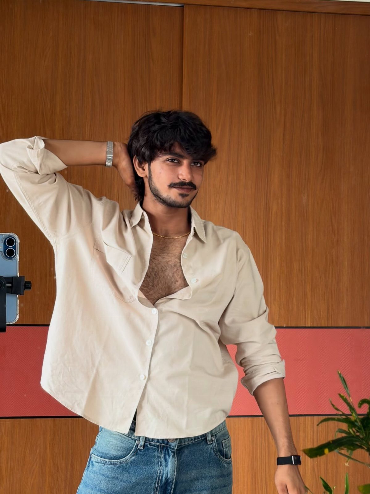 Man in a cream shirt and blue jeans posing with one hand behind his head.