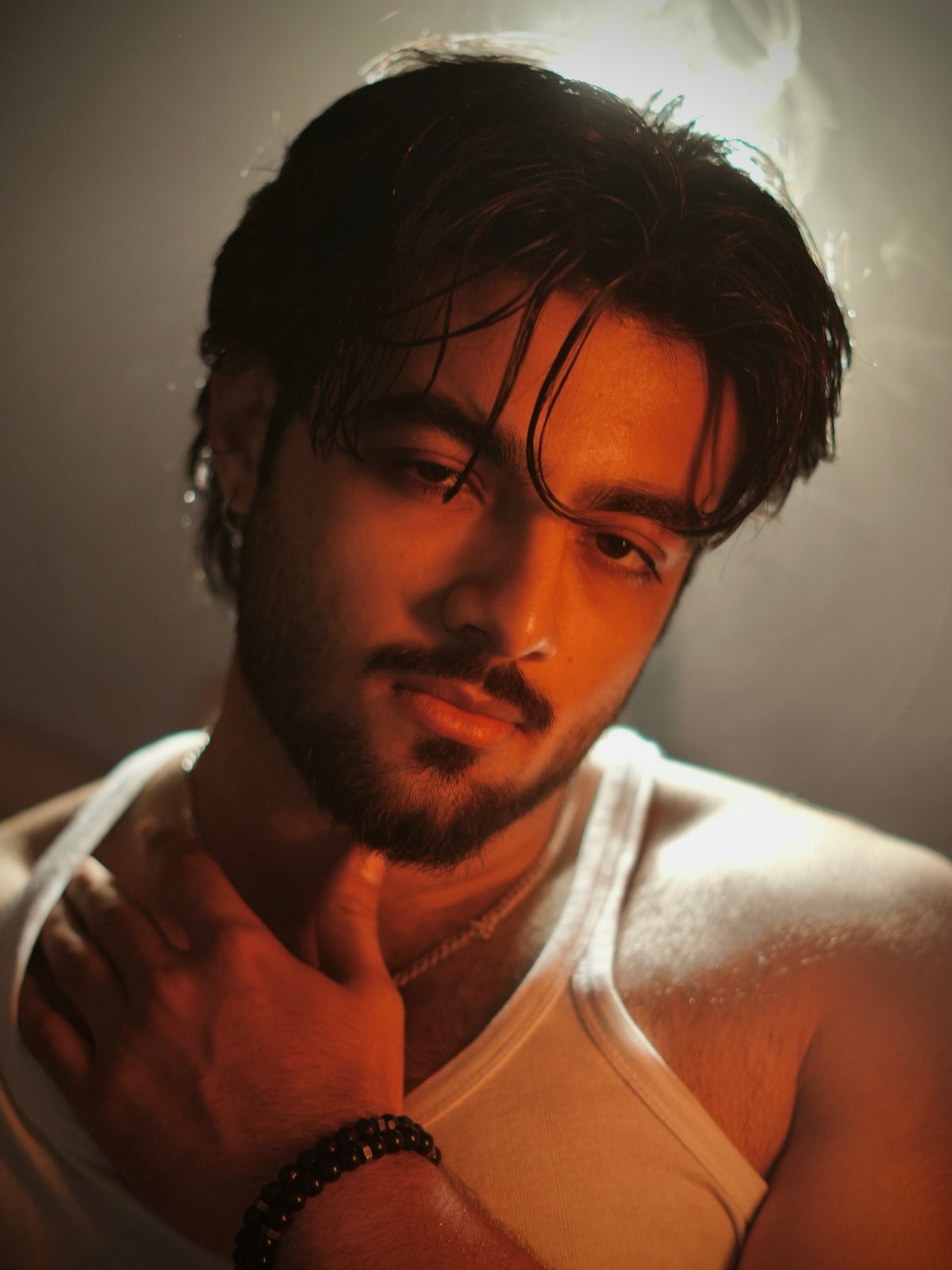 A young man with dark hair and a beard posing in a white tank top under warm light.