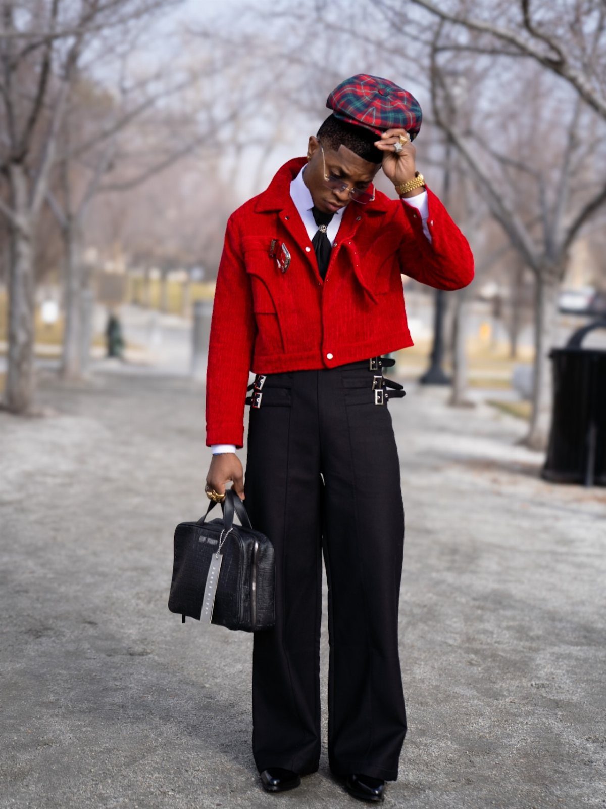 Man in a red jacket and black trousers standing on a path while touching his plaid cap.