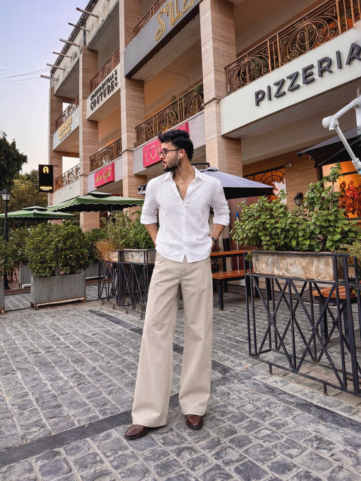 Man in white shirt and beige trousers standing on a cobblestone street by a pizzeria.
