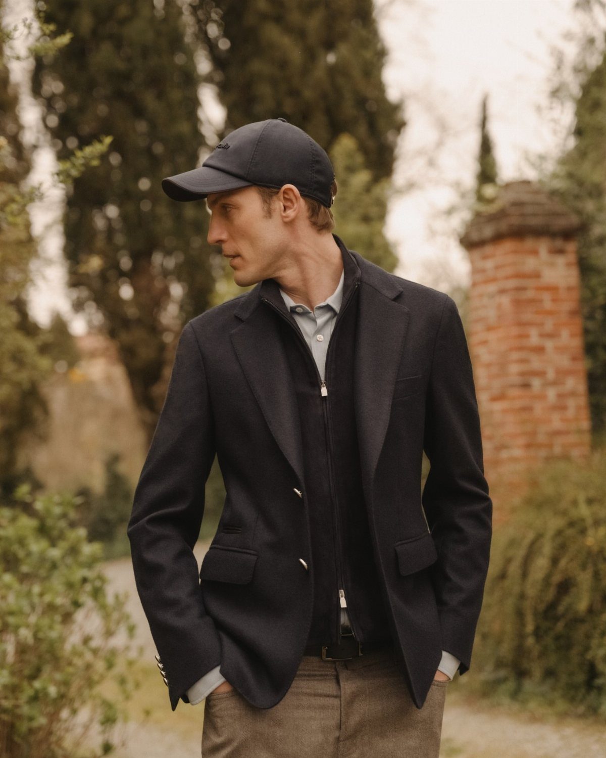 Man in a dark blazer and baseball cap standing with hands in his pockets outdoors.