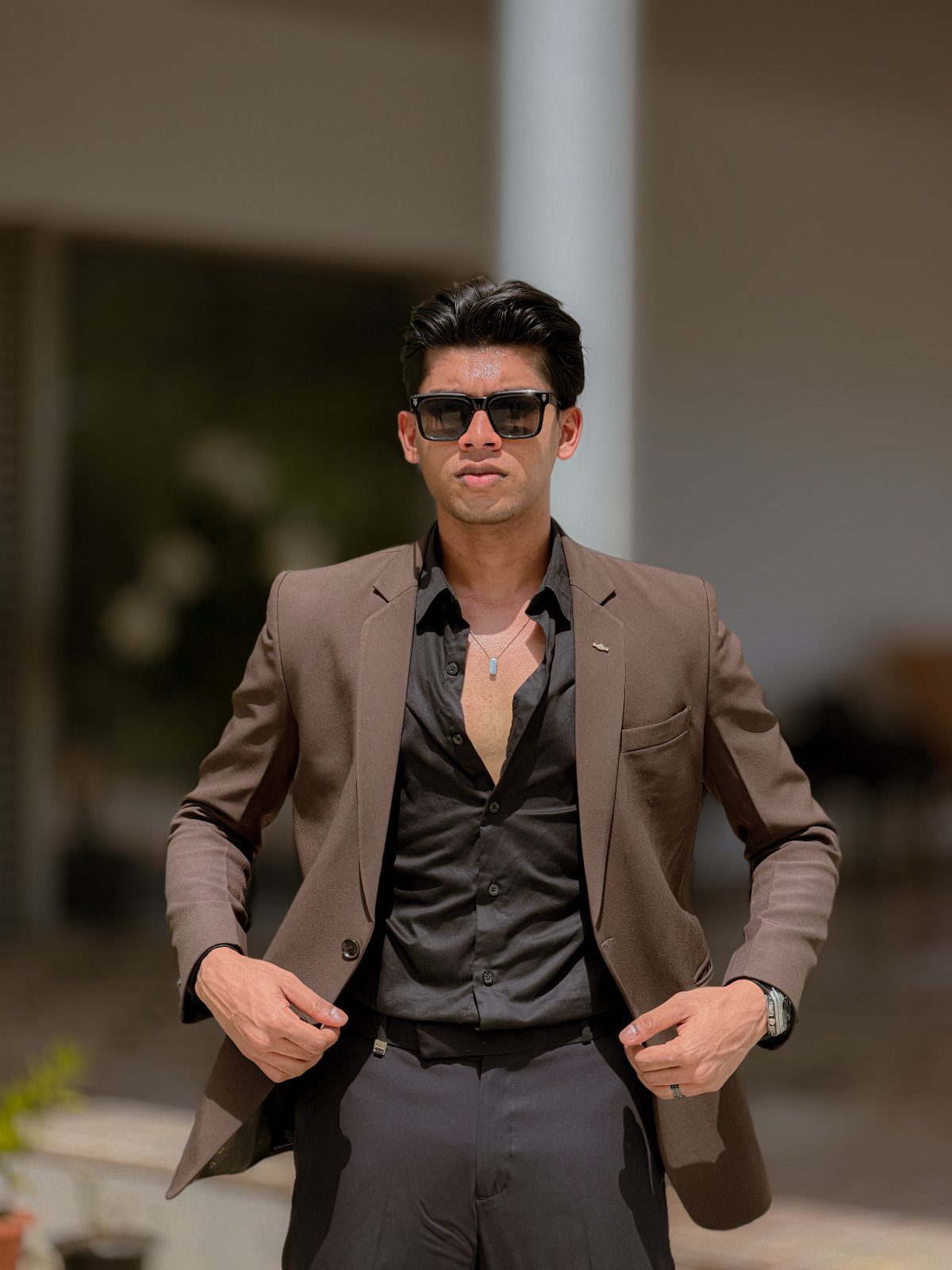 Man in a brown blazer and sunglasses posing while holding his jacket edges.