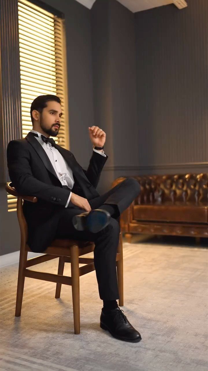 Man in a black tuxedo sitting on a wooden chair with legs crossed.