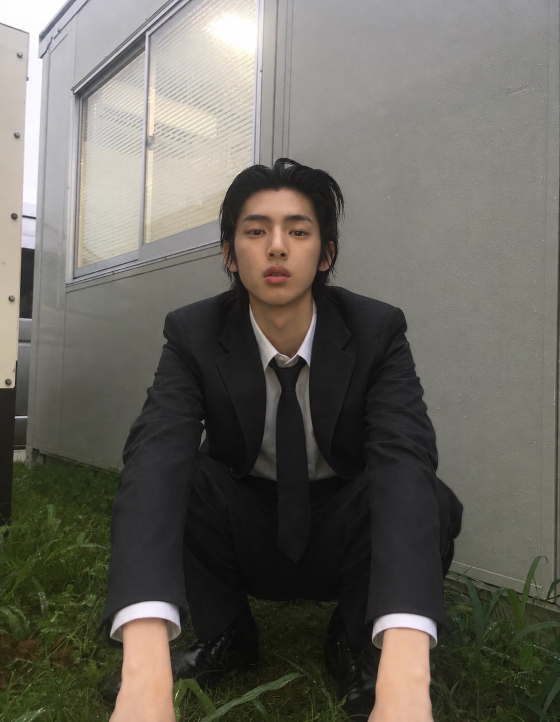 A man in a black suit squatting on green grass in front of a building.