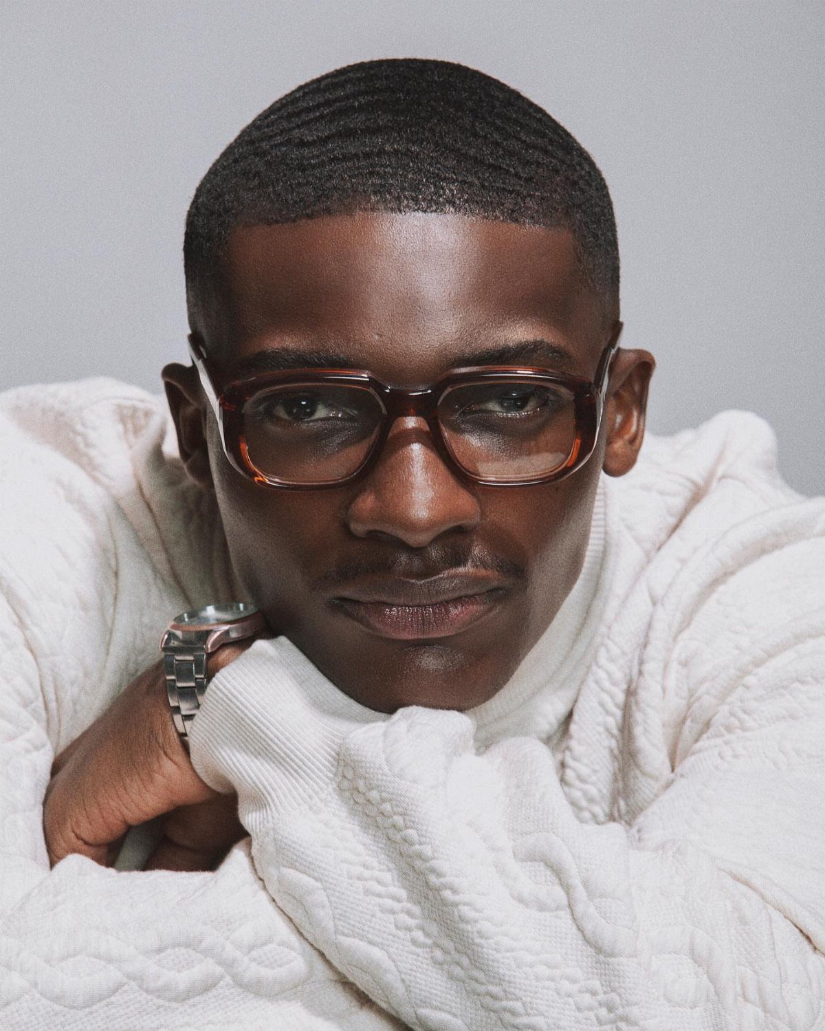 A man with glasses and a white sweater resting his chin on his hands.