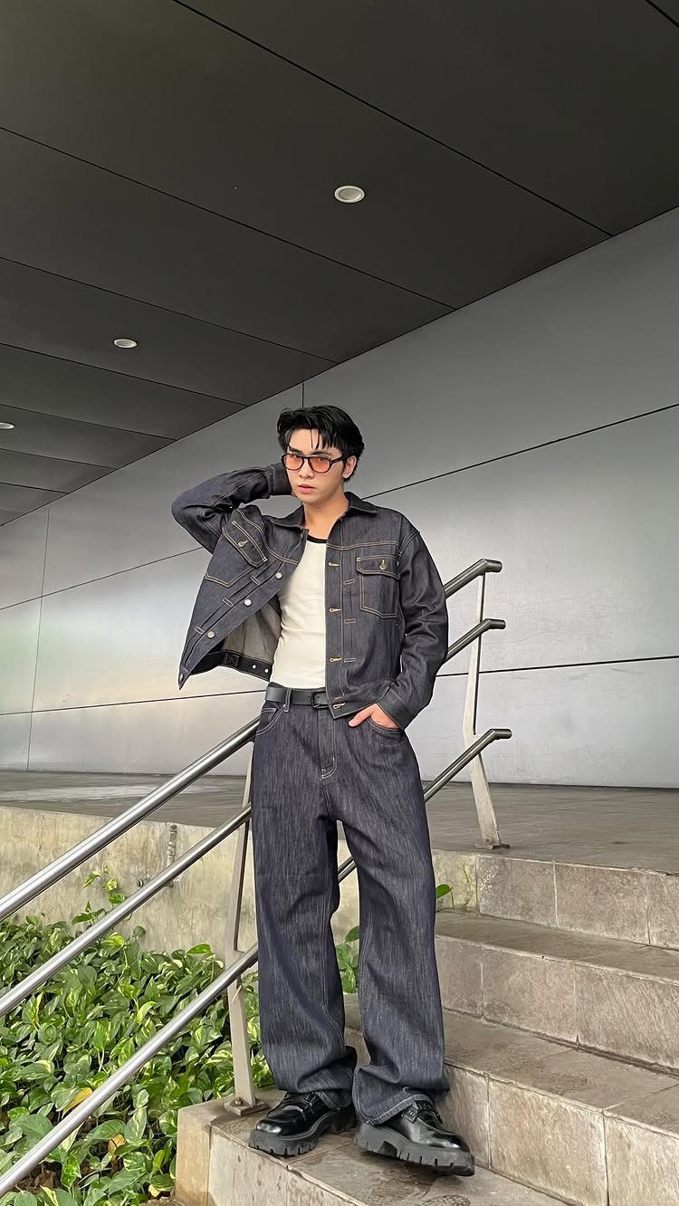 A young man in denim clothing posing on concrete stairs next to a metal handrail.