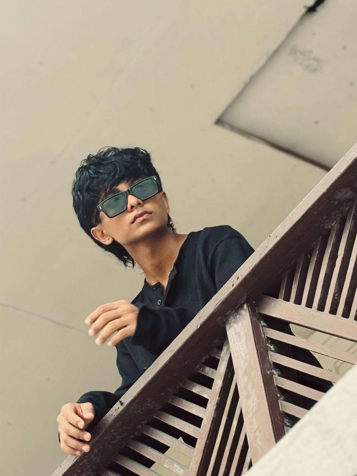 Young man in a black shirt wearing sunglasses, posing behind a wooden railing.