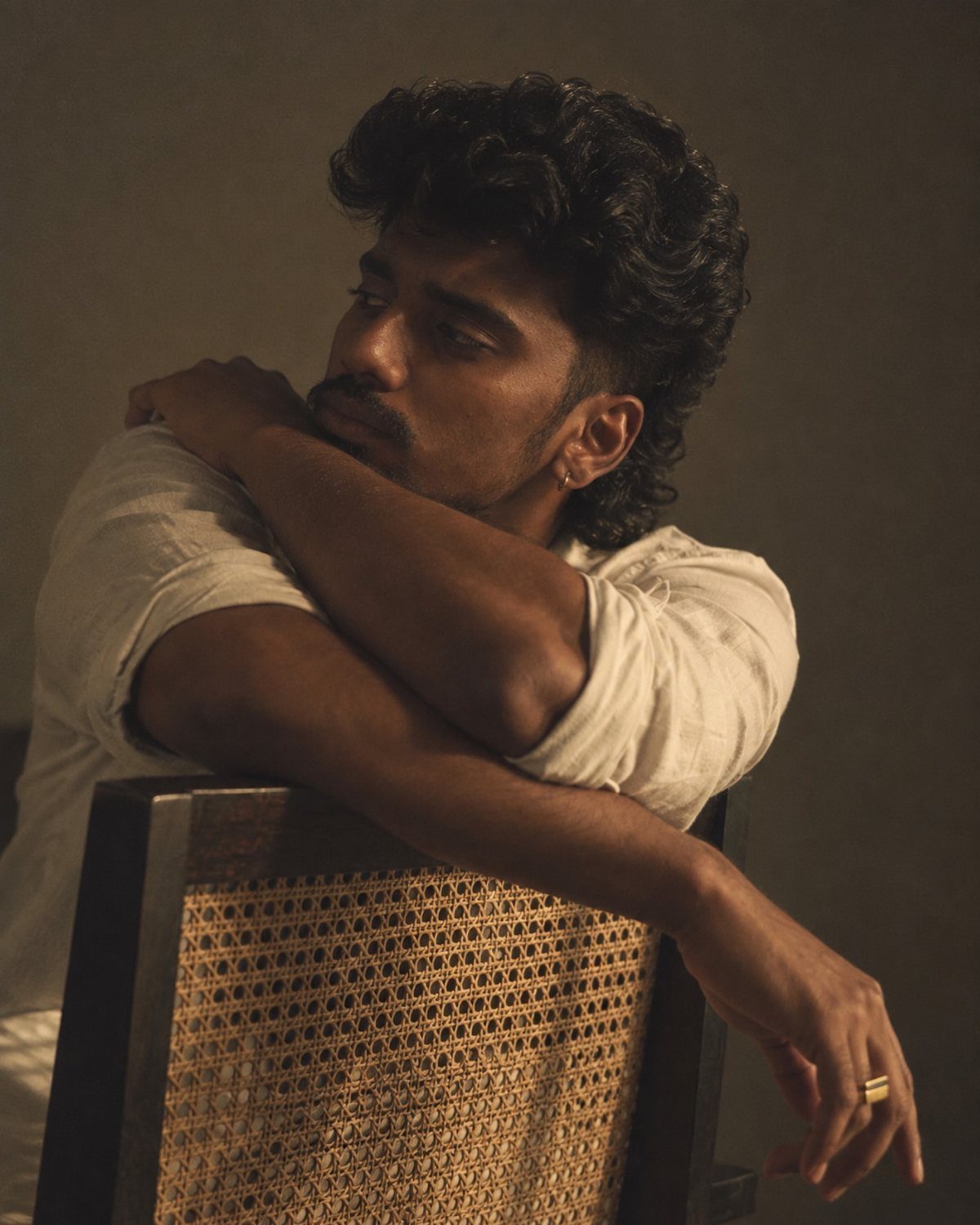 A thoughtful young man with curly hair resting his arms on a chair back while looking away.