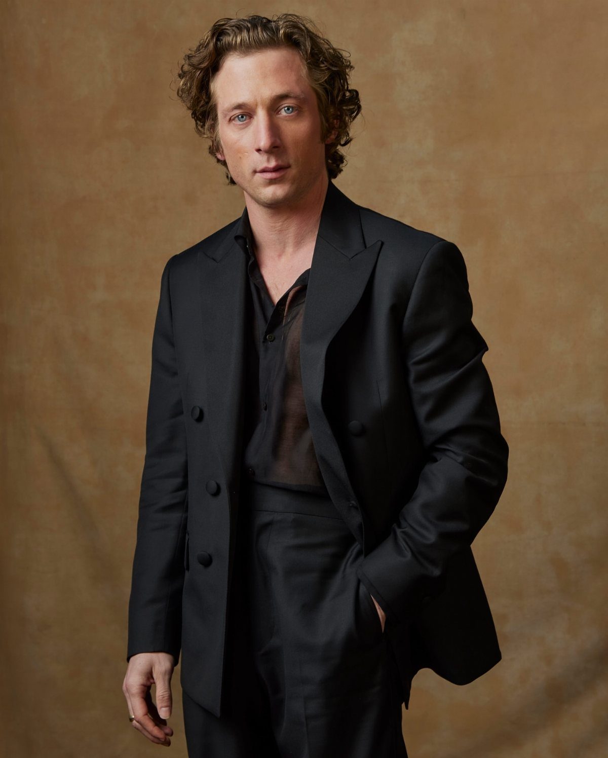 A man wearing a black blazer and a black sheer button-down shirt against a tan background.