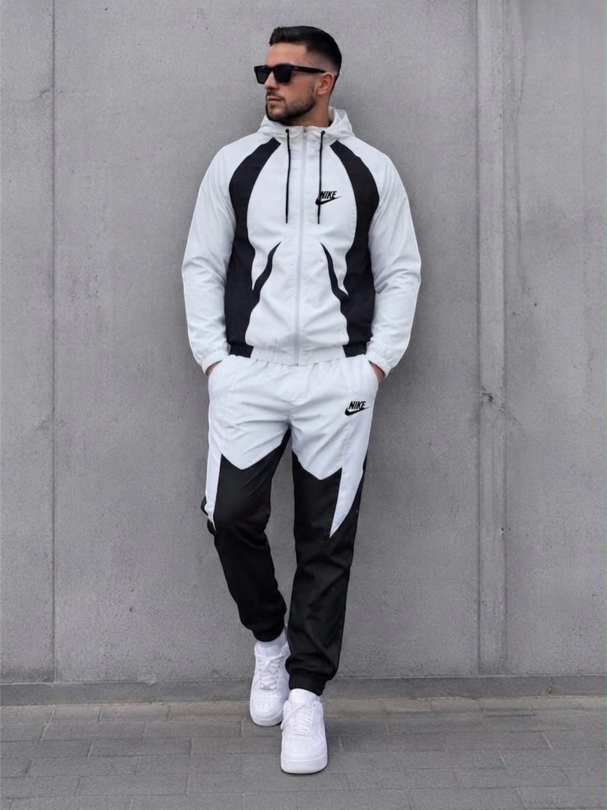 Man in a white and black Nike tracksuit and sunglasses standing against a concrete wall.