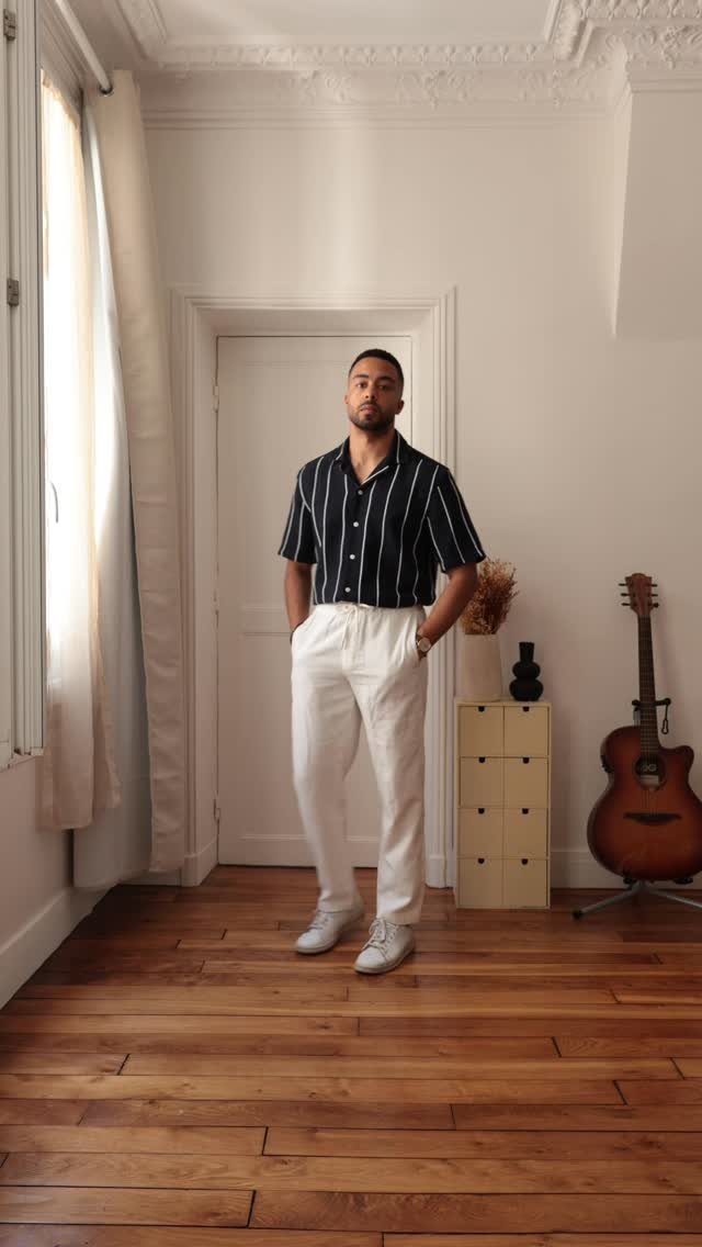 Man wearing a striped Cuban collar shirt and white trousers standing in a bright room.