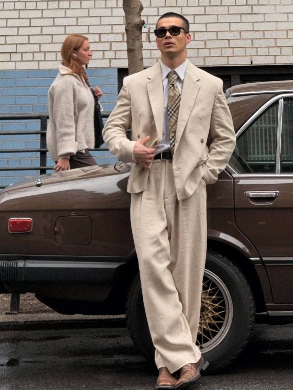 A man in a cream linen suit and sunglasses leaning against a brown car.
