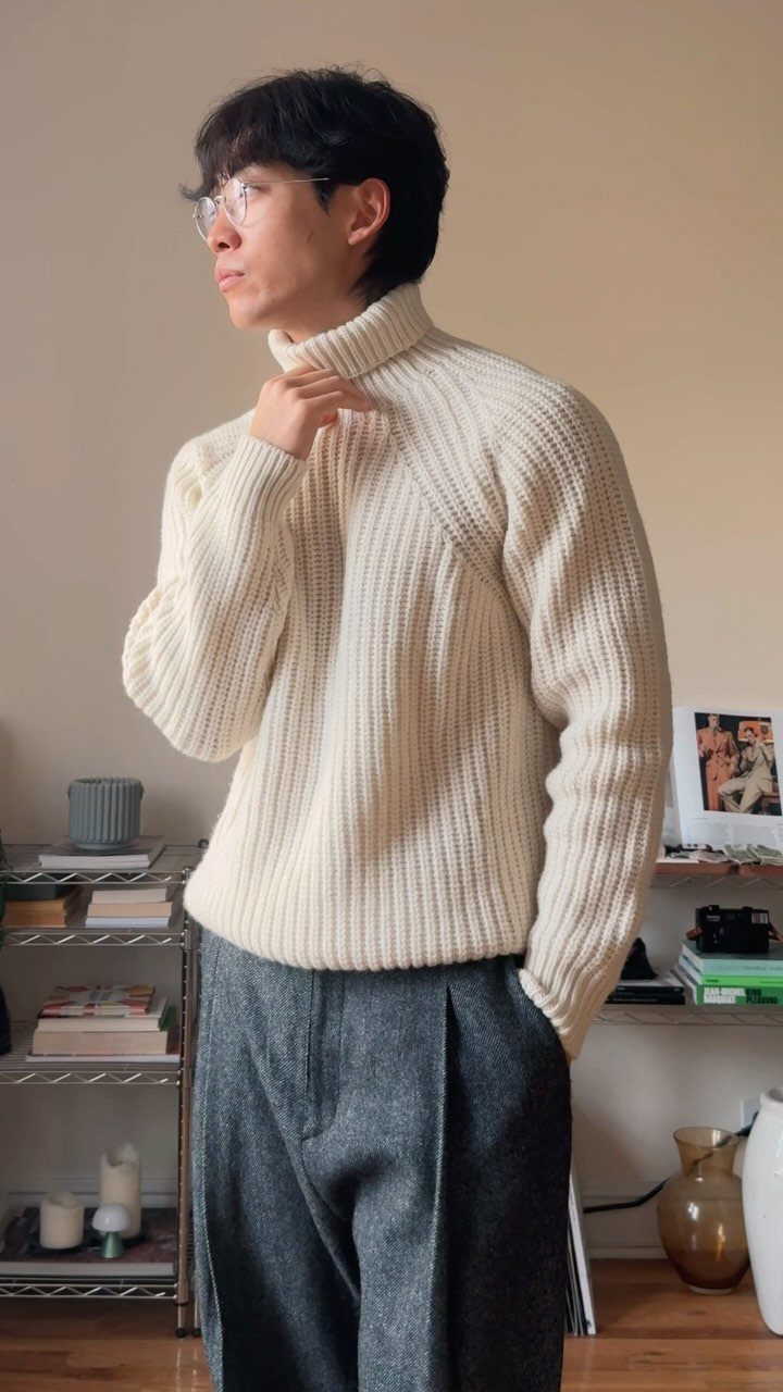 Man in a cream ribbed turtleneck sweater and grey trousers standing in a room.