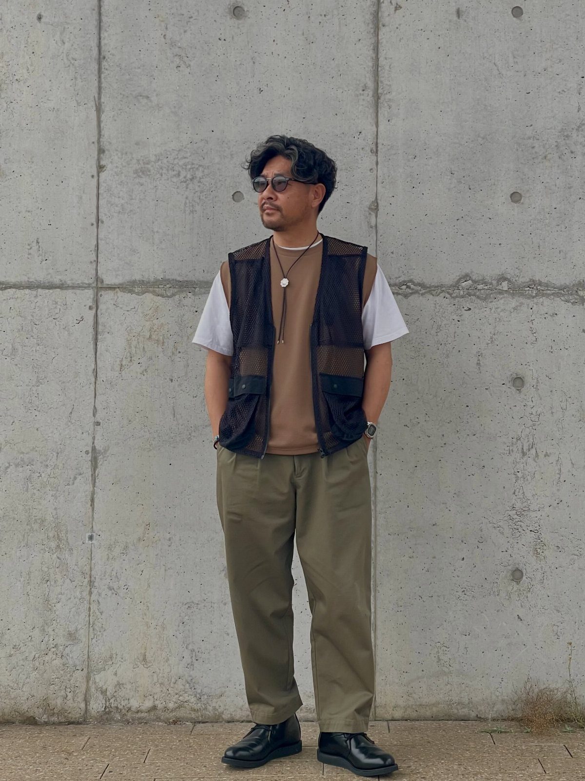A man posing in a dark patterned utility vest, brown t-shirt, and olive trousers against a concrete wall.