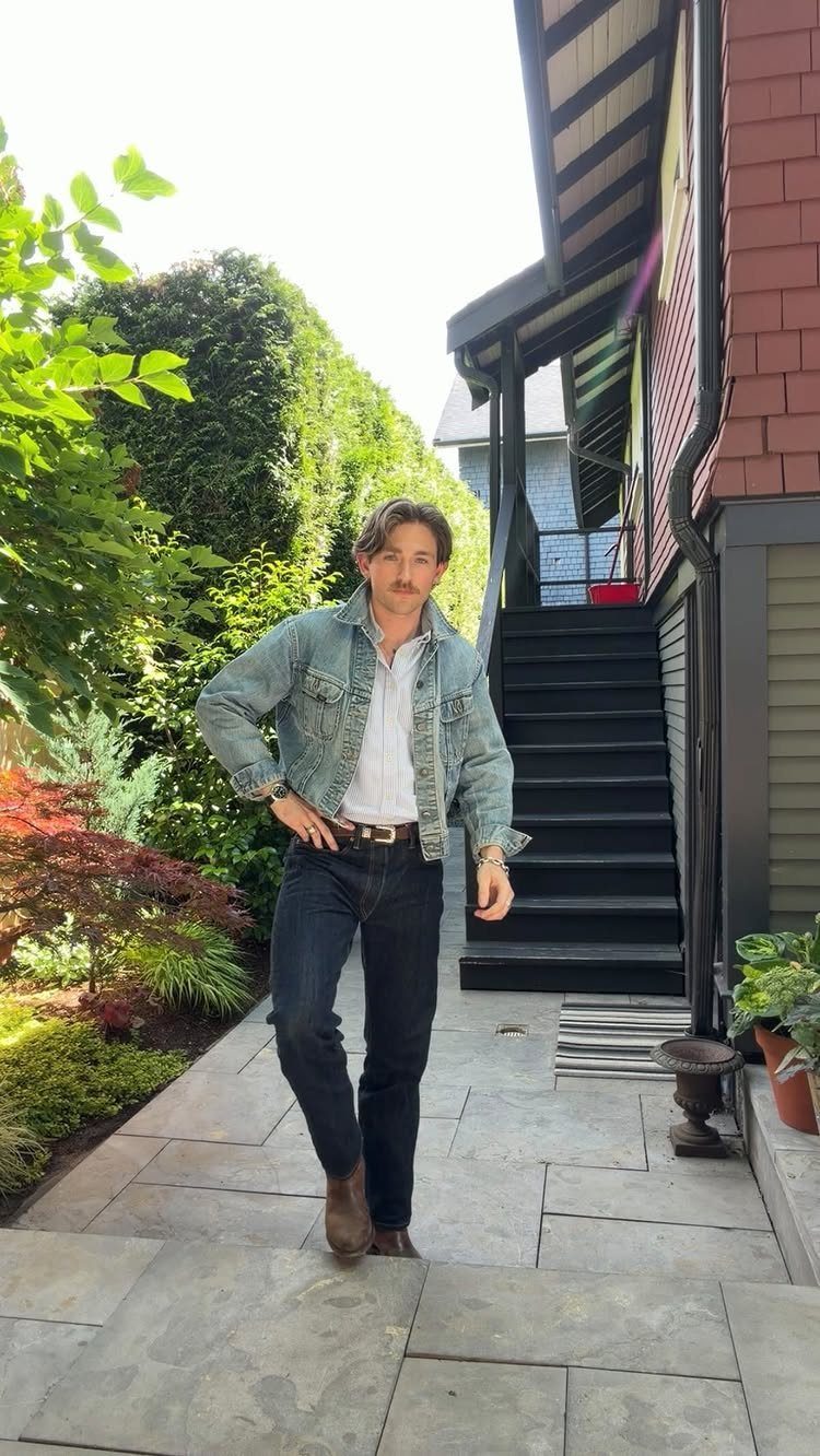 Man in a denim jacket and dark jeans posing while wearing brown leather boots outdoors.
