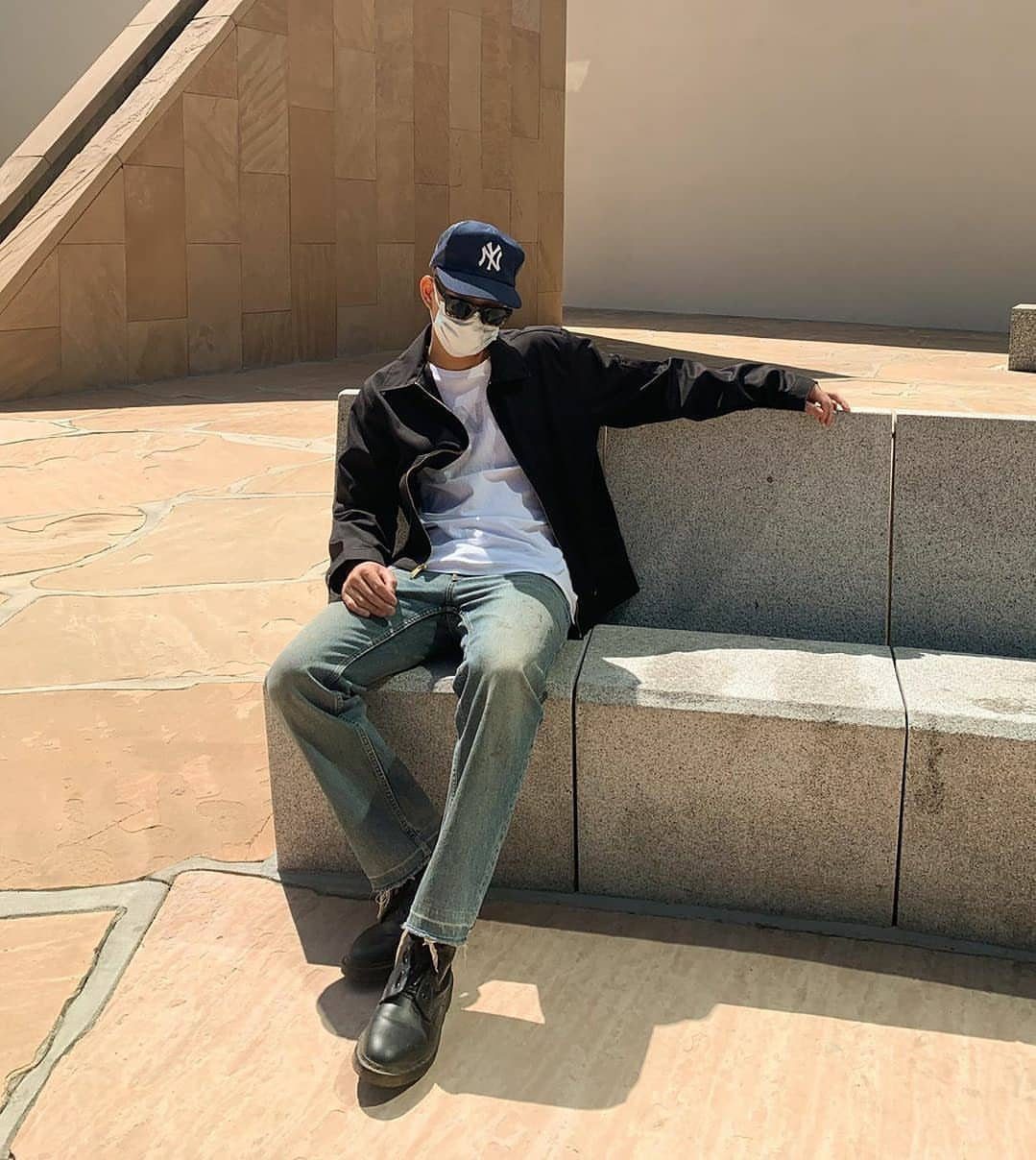 Man in a black jacket and blue jeans sitting on a stone ledge wearing black combat boots.