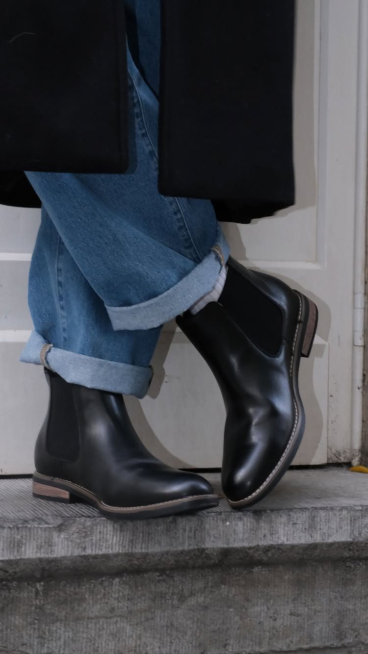 Black leather Chelsea boots worn with cuffed blue jeans and a black coat.