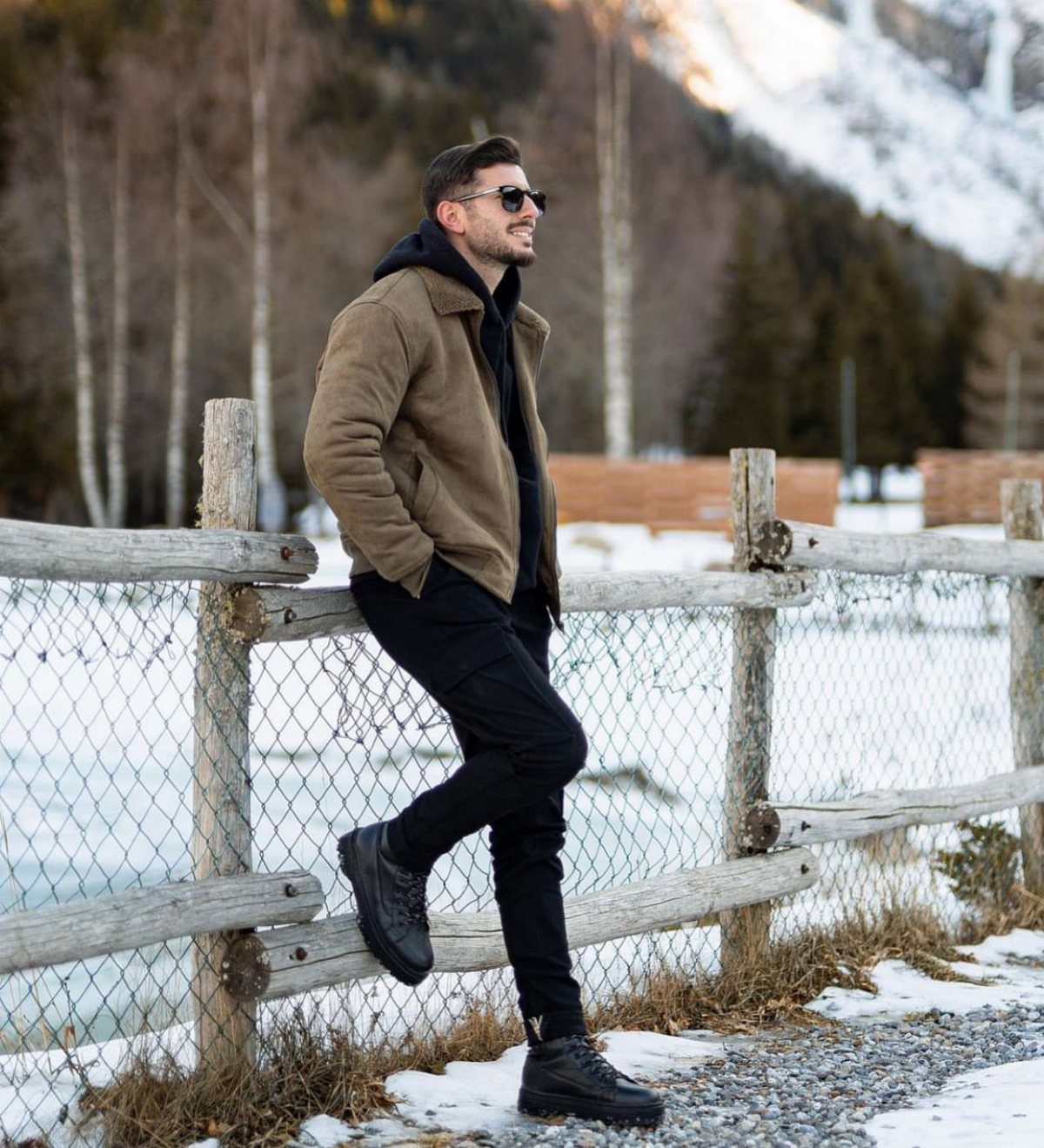 Man in a brown jacket and black boots leaning against a wooden fence in the snow.