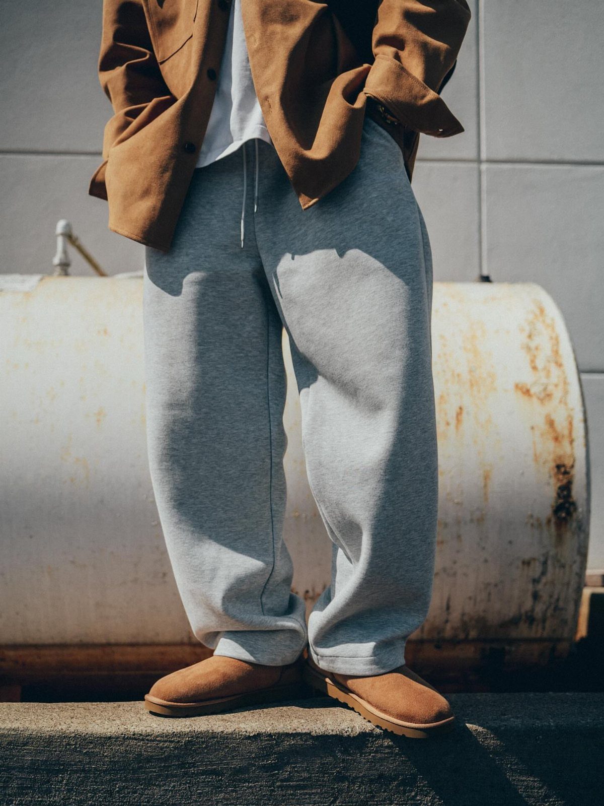 Person wearing a brown shirt, grey sweatpants, and brown suede boots standing by a metal tank.