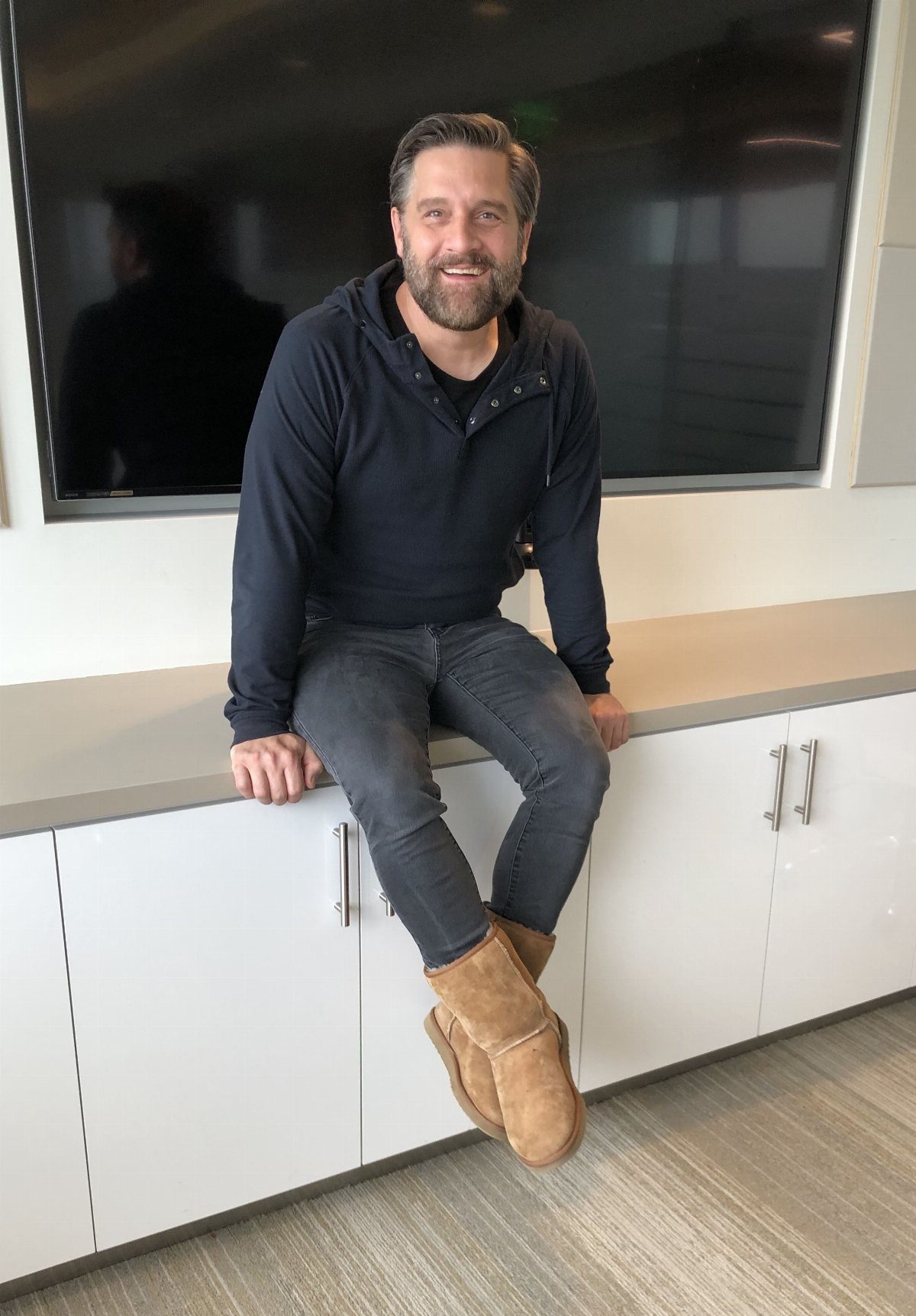 Man in navy hoodie, grey jeans, and tan Ugg boots sitting on a white cabinet.