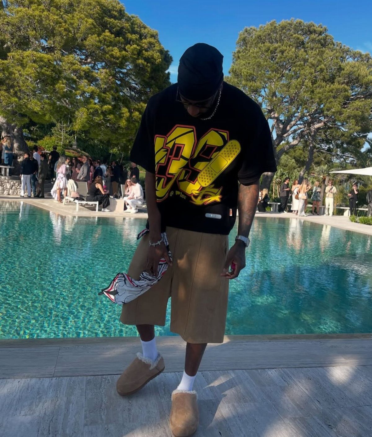 Man in a black graphic t-shirt and tan shorts wearing Ugg slides by a pool.