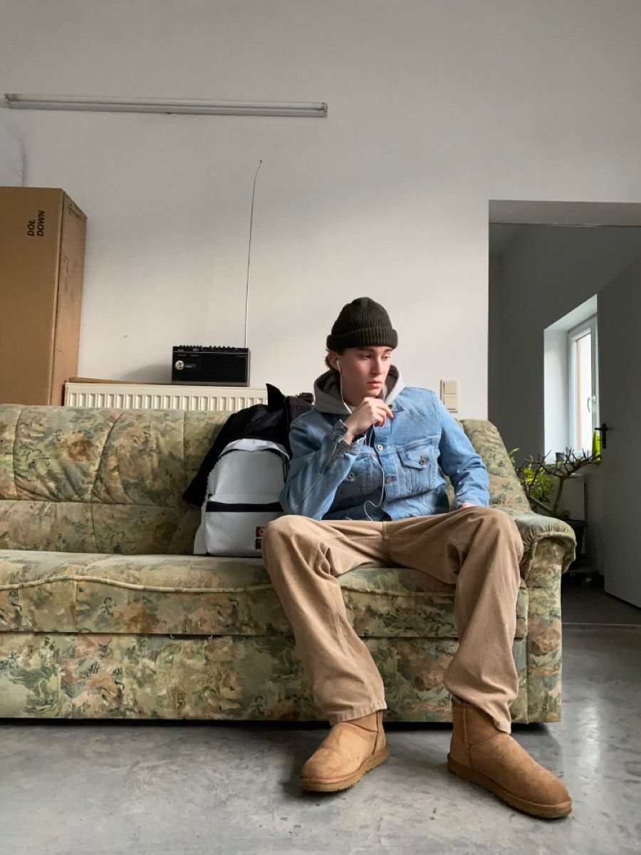 Man sitting on a floral sofa wearing a denim jacket, tan pants, and tan boots.