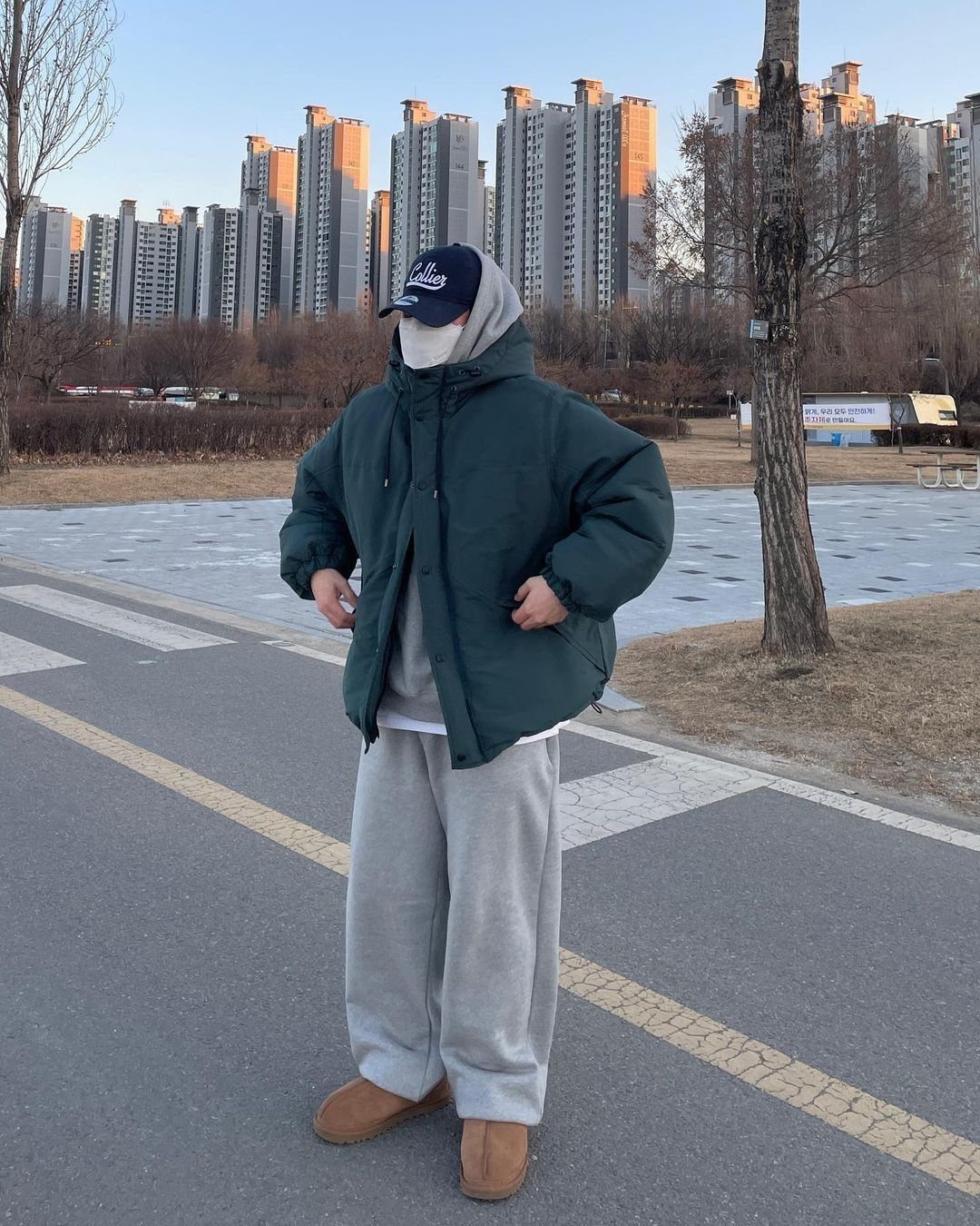 A person wearing a green puffer jacket, grey sweats, and tan boots standing outdoors.