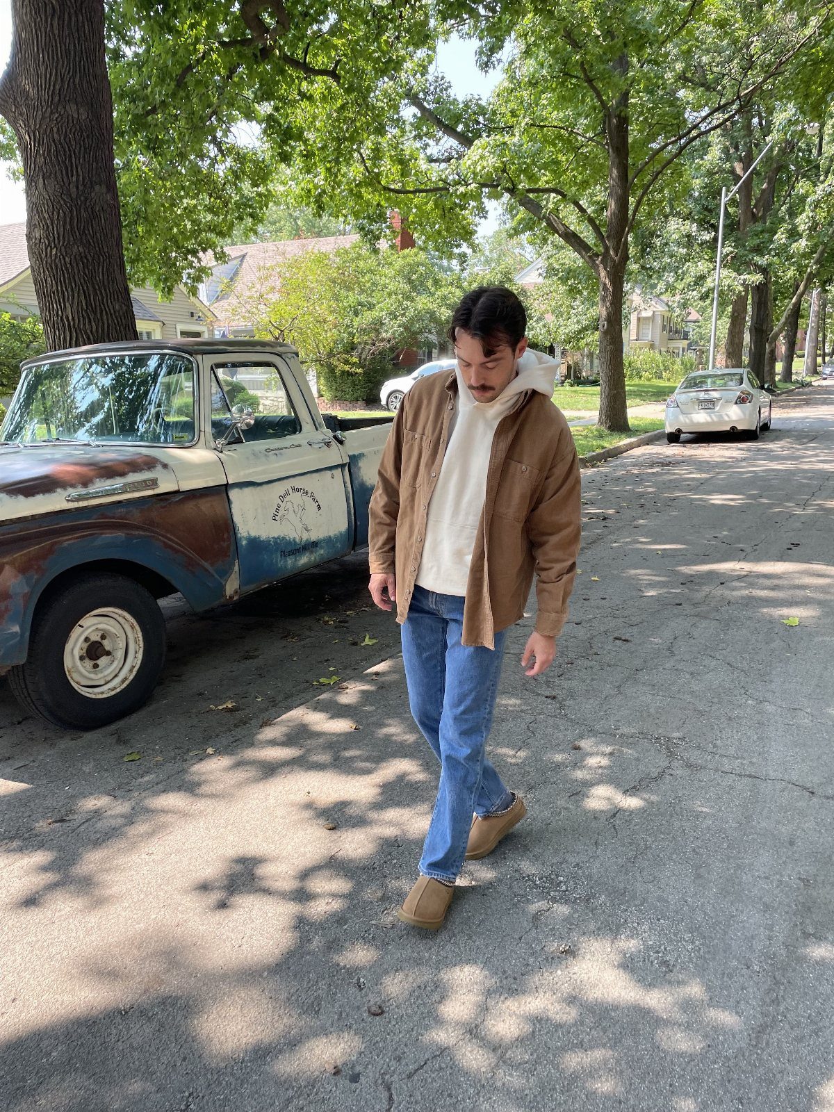 Man walking on a street wearing a brown jacket, white hoodie, blue jeans, and tan Ugg boots.
