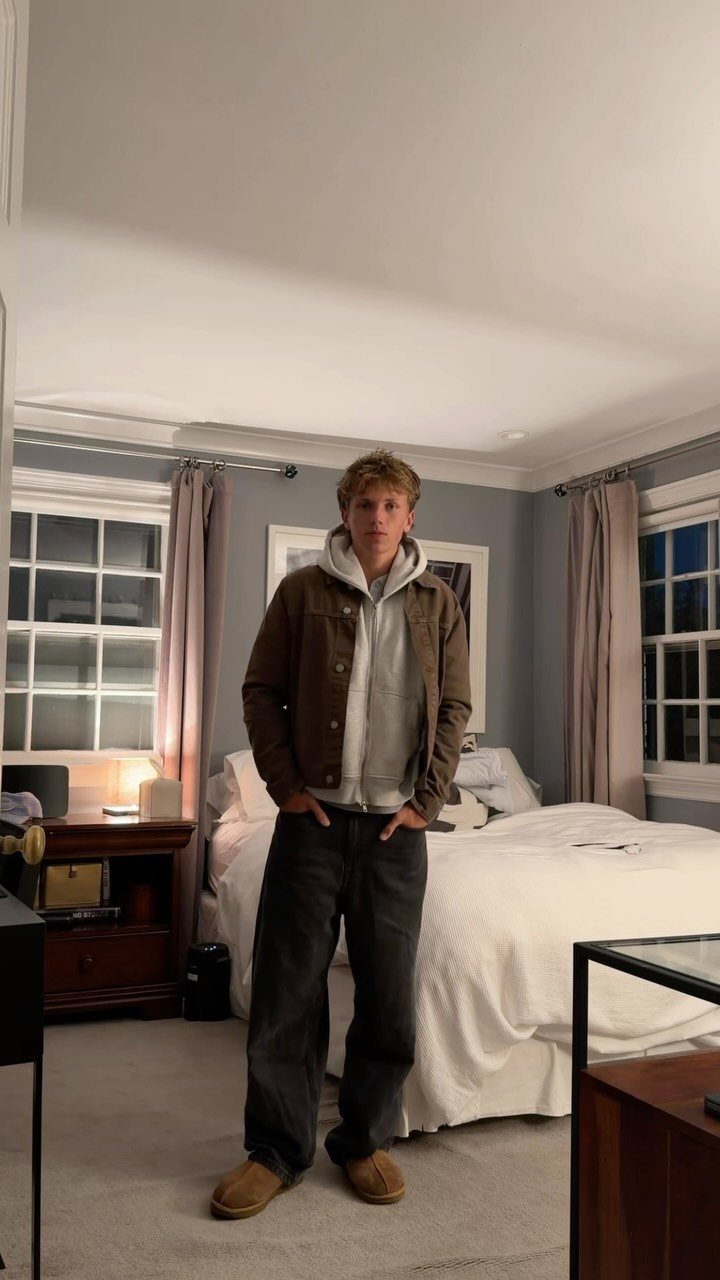 Young man posing in a bedroom wearing a brown jacket, grey hoodie, dark pants, and tan Ugg boots.