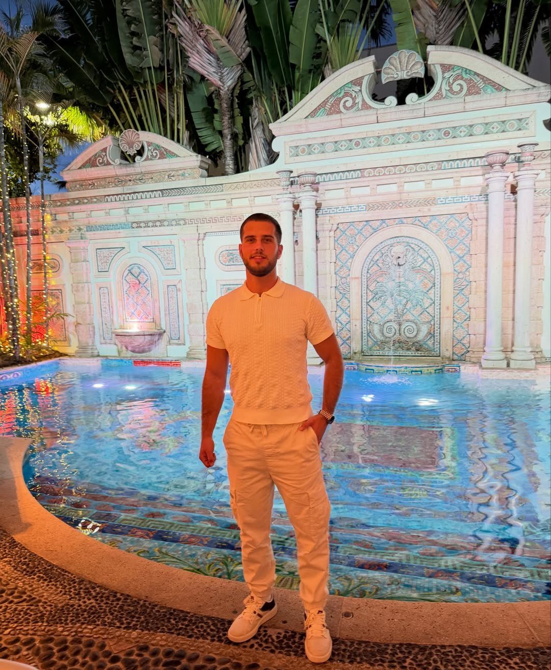 Man in an all-white polo shirt and cargo trousers standing by a swimming pool.