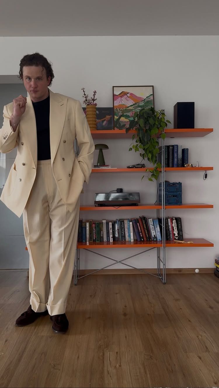 Man in a cream double-breasted suit standing in a room with orange bookshelves.