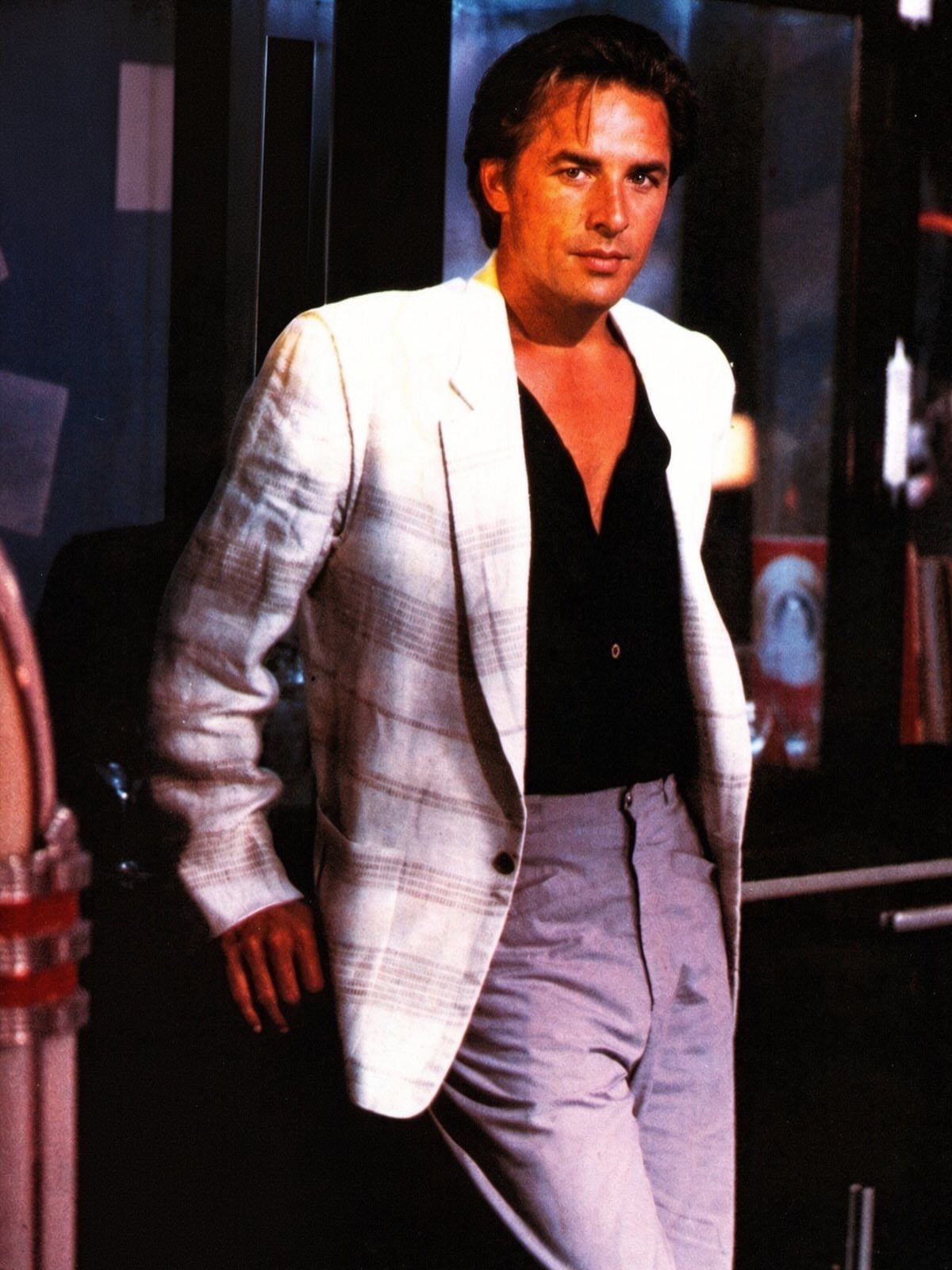 Man in a white plaid blazer, black shirt, and light grey trousers posing indoors.