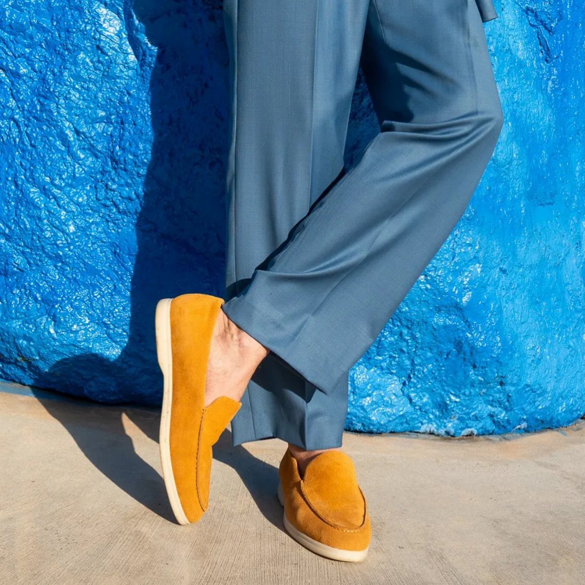 Light blue trousers and mustard yellow suede loafers against a bright blue textured wall.