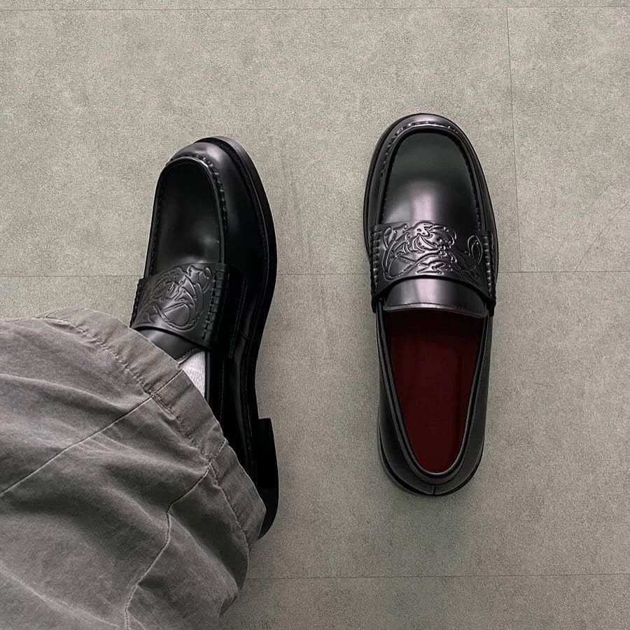 Black leather loafers with embossed patterns on a grey tiled floor next to grey trousers.
