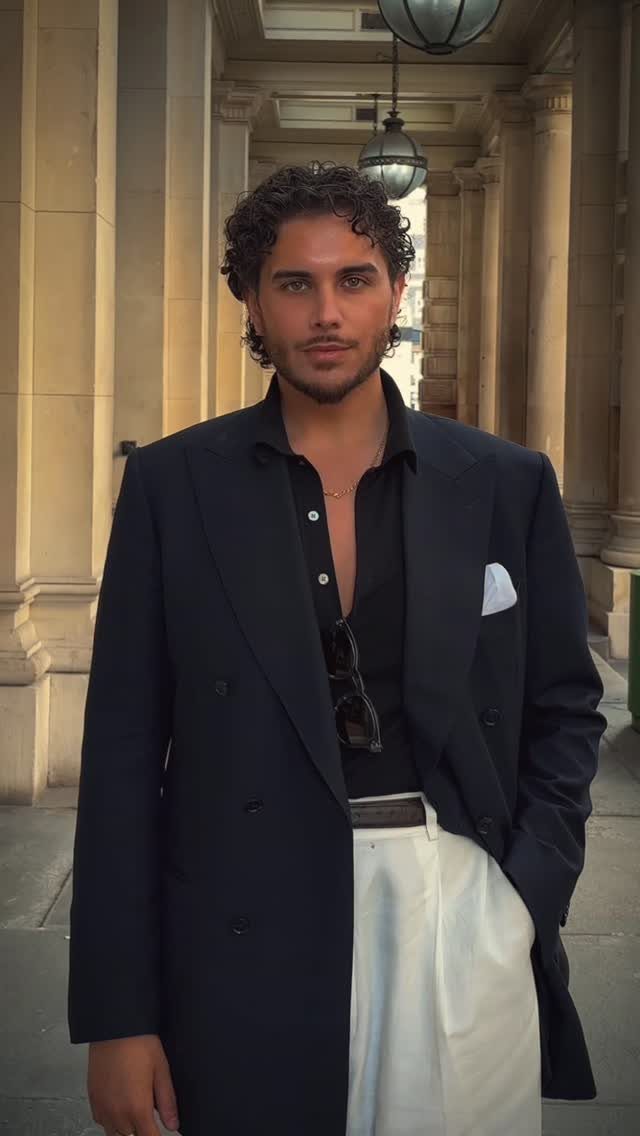 Man with curly hair wearing a navy blazer and white trousers standing in a stone corridor.