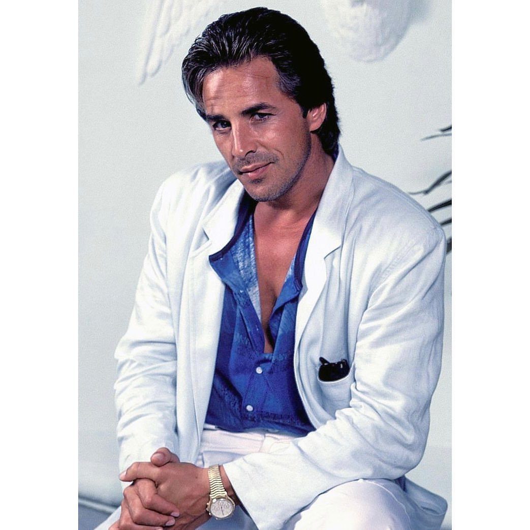A man wearing a white unconstructed jacket and a blue patterned shirt while posing.