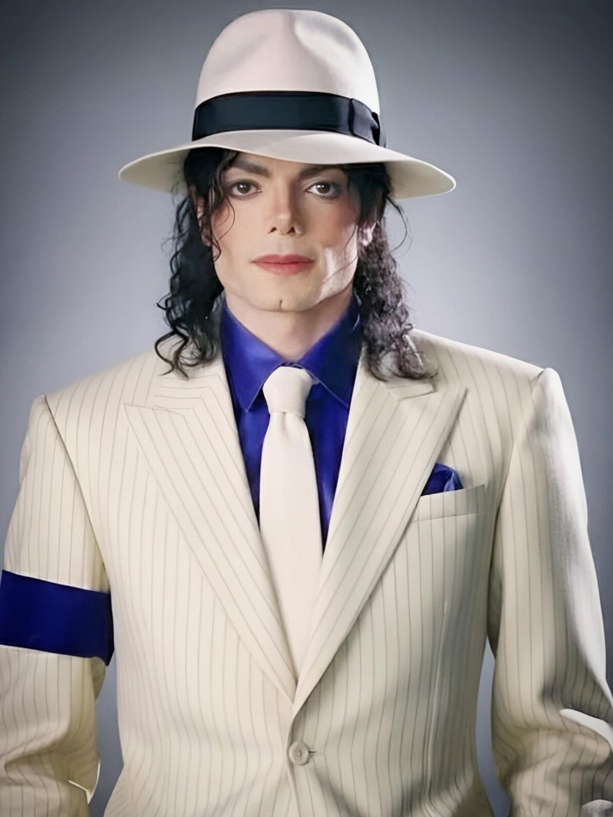 Man in a white pinstripe suit, blue shirt, white tie, and a blue armband on his sleeve.