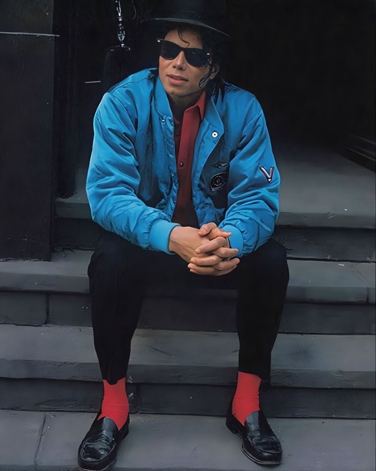 Michael Jackson sitting on steps wearing a blue jacket, red shirt, black trousers, and red socks.