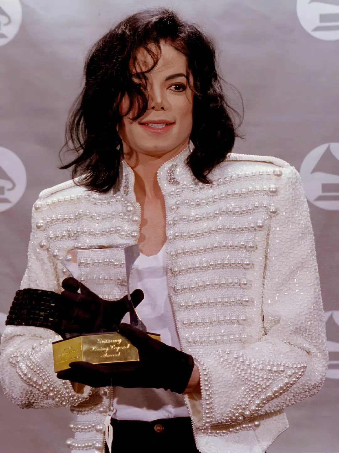 Michael Jackson in a white pearl-embellished military jacket holding a gold award.