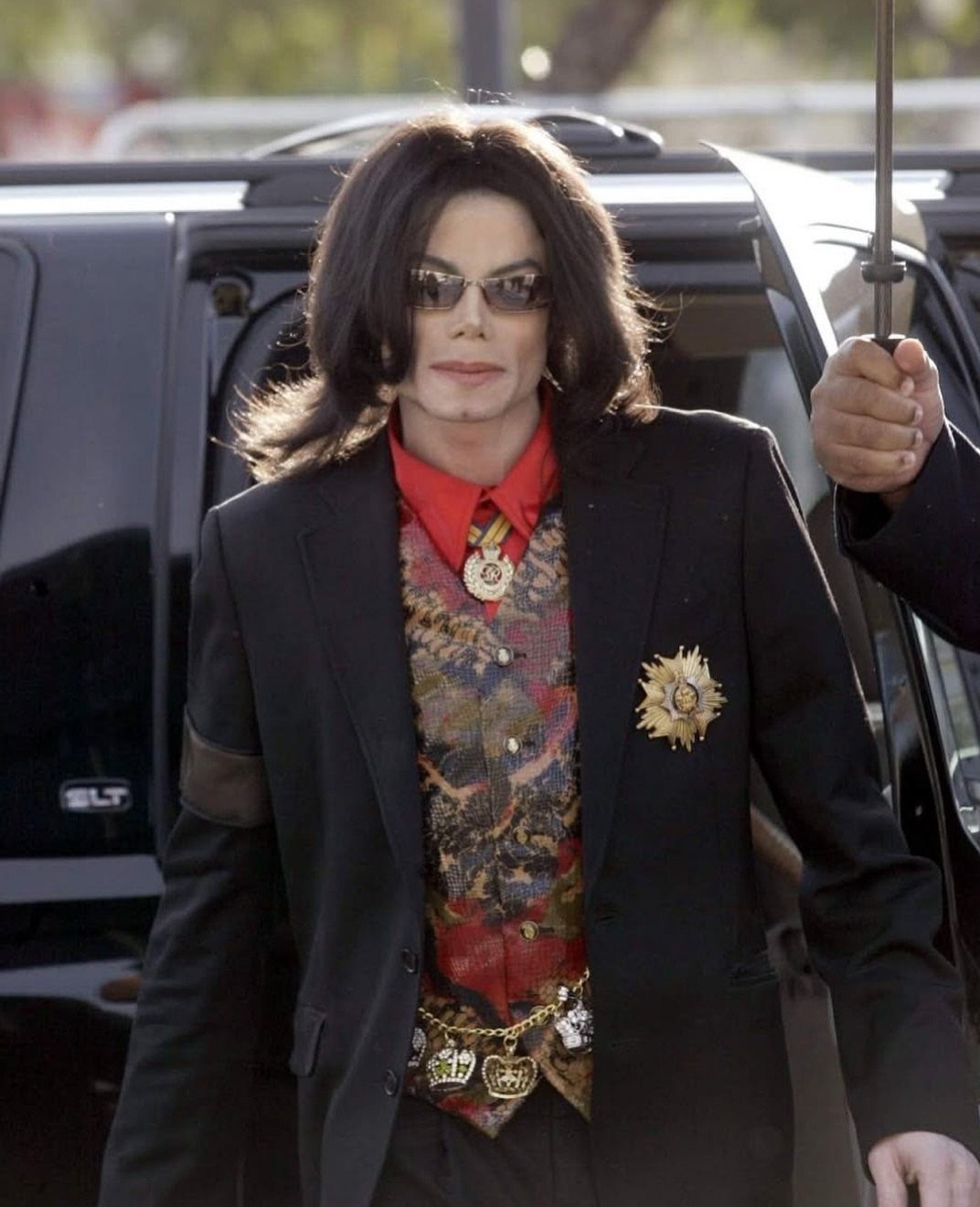 Michael Jackson wearing a black blazer, patterned vest, and red shirt while standing near a vehicle.