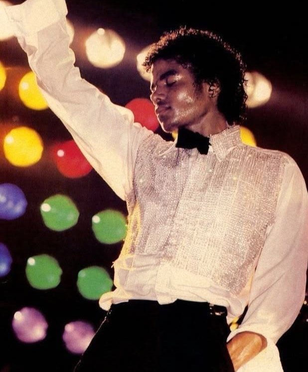 Michael Jackson in a white sequined shirt and black bow tie against colorful bokeh lights.