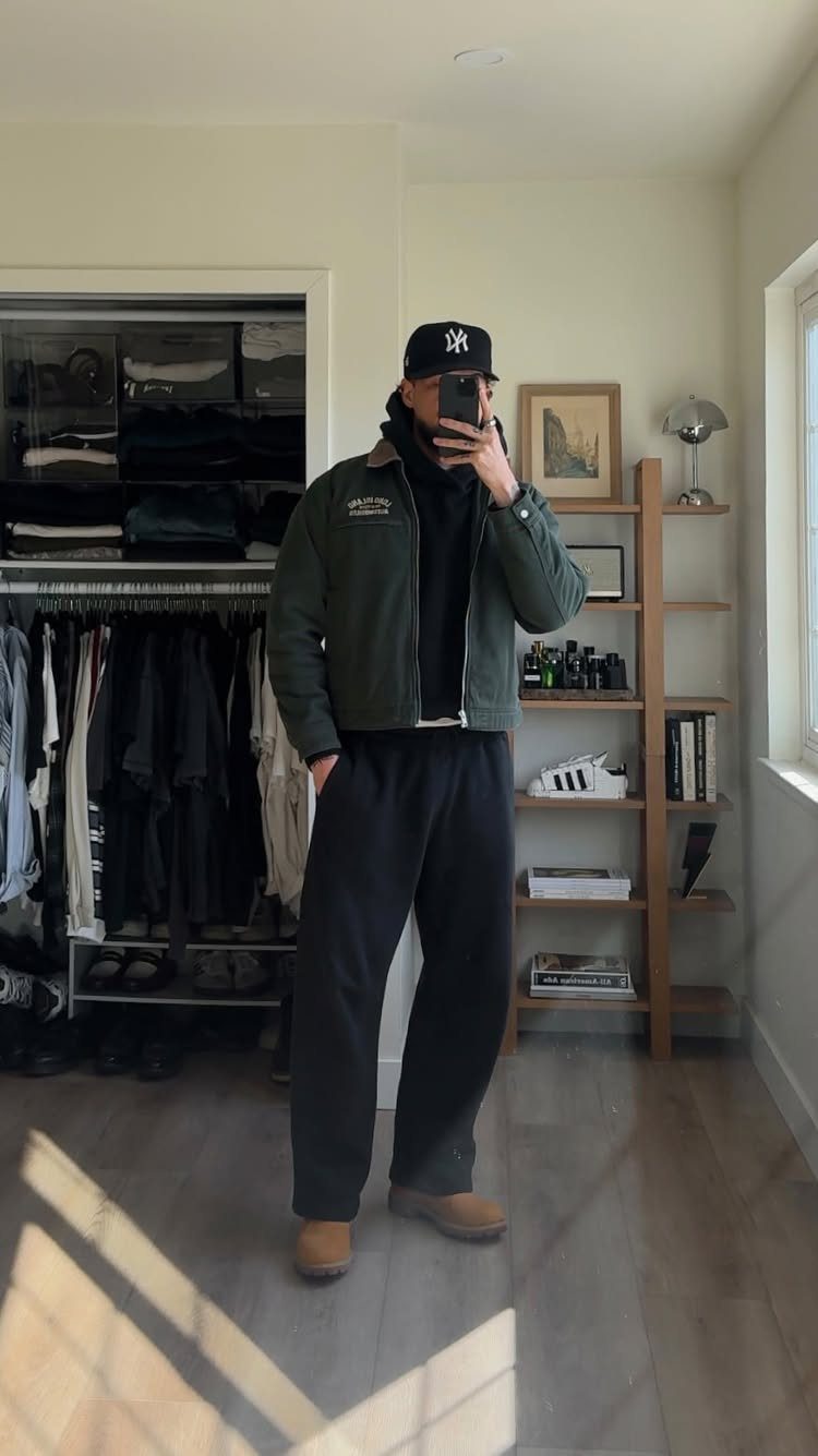 Man in olive bomber jacket, black trousers, and tan boots posing for a mirror selfie.