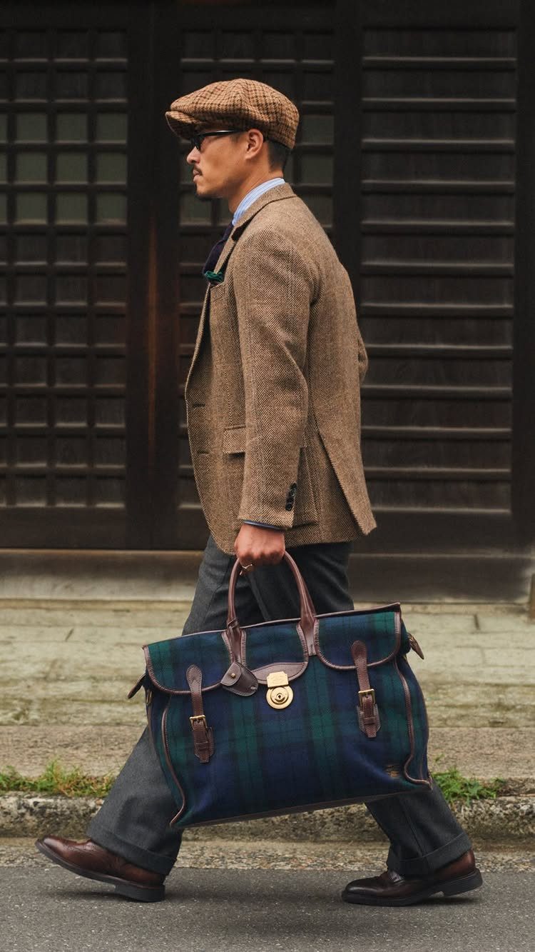 A man in a tweed blazer and flat cap walking while carrying a large plaid bag.