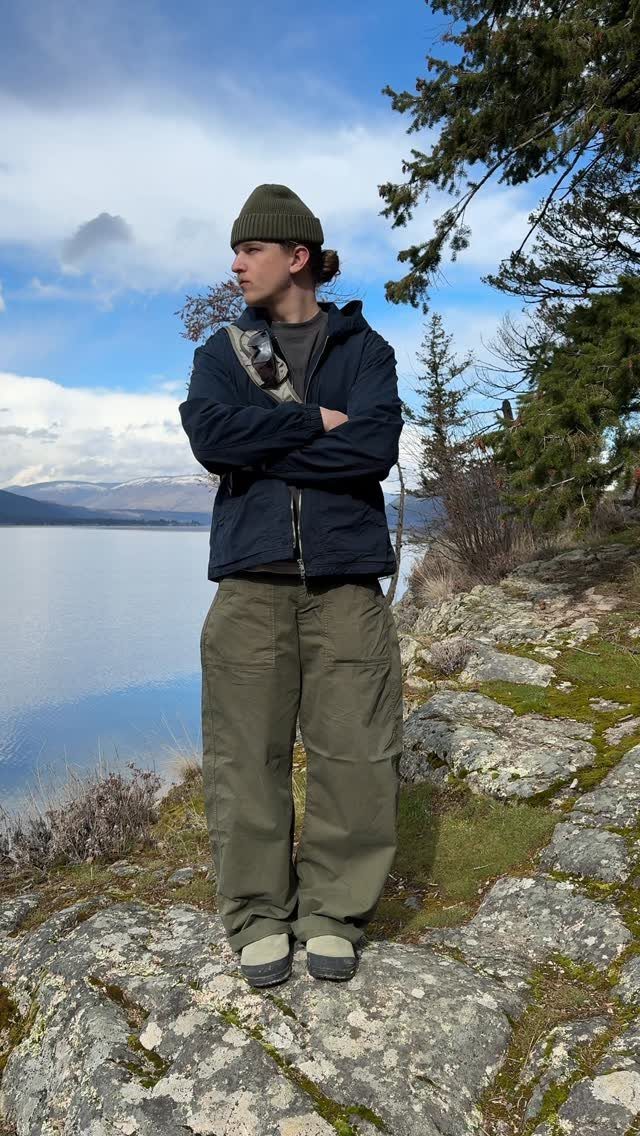 Man in technical outdoor clothing standing on rocks by a large lake under a blue sky.